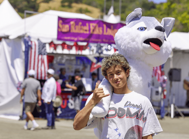 A teenager holding a large wolf stuffed animal on his shoulder in front of a county fair tent.