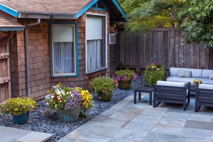 Well-maintained and potted flowers in between a brown house and a stone patio.