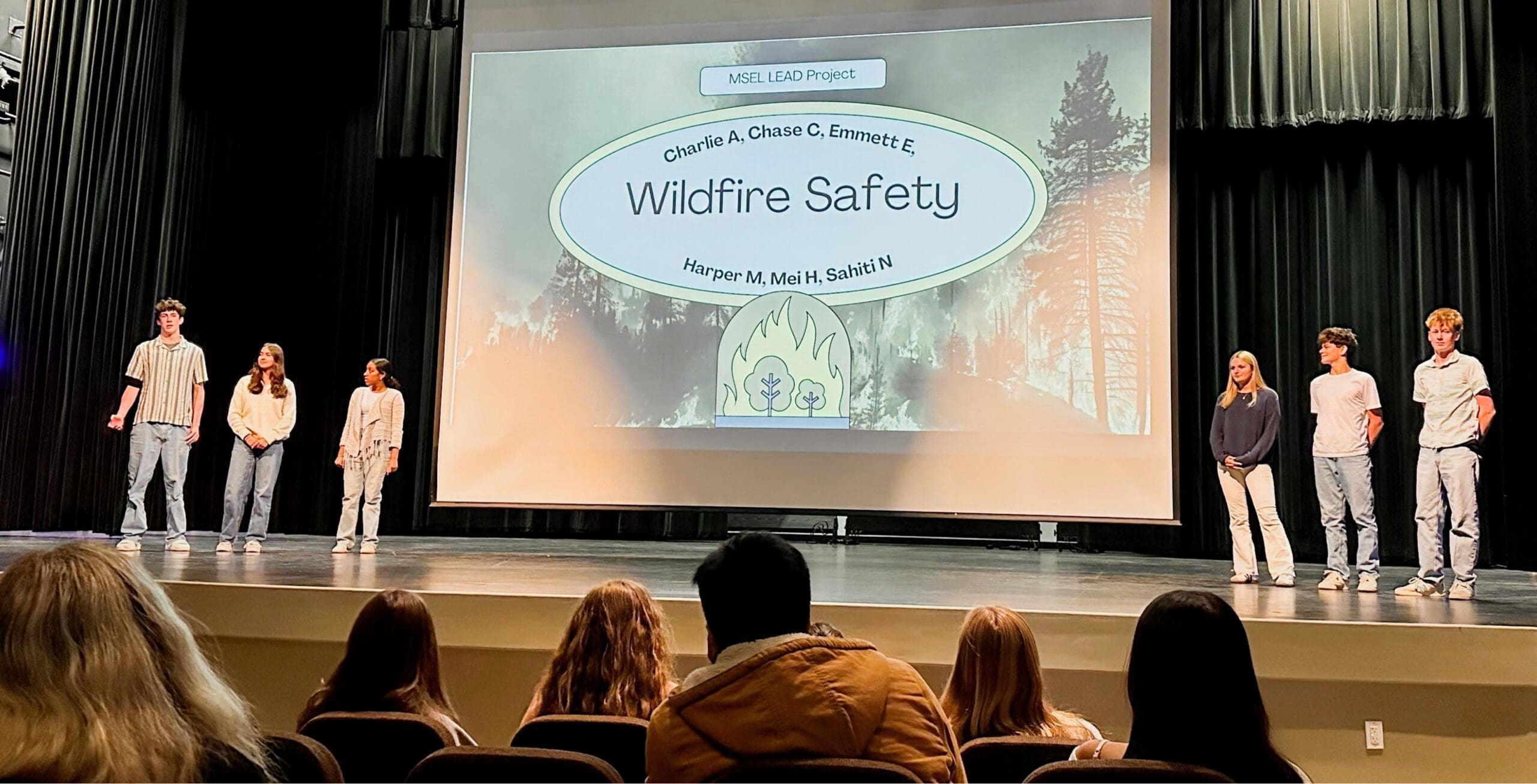 A group of MSEL students on a stage with a projector screen, giving their final presentation to a diverse crowd of people.
