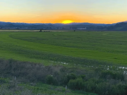 A horizontal view of a large, green open space with the sun setting behind mountains in the background.