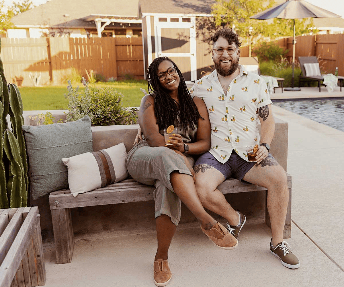 A young couple on a bench besides a pool with drinks in their hands, smiling at the camera.